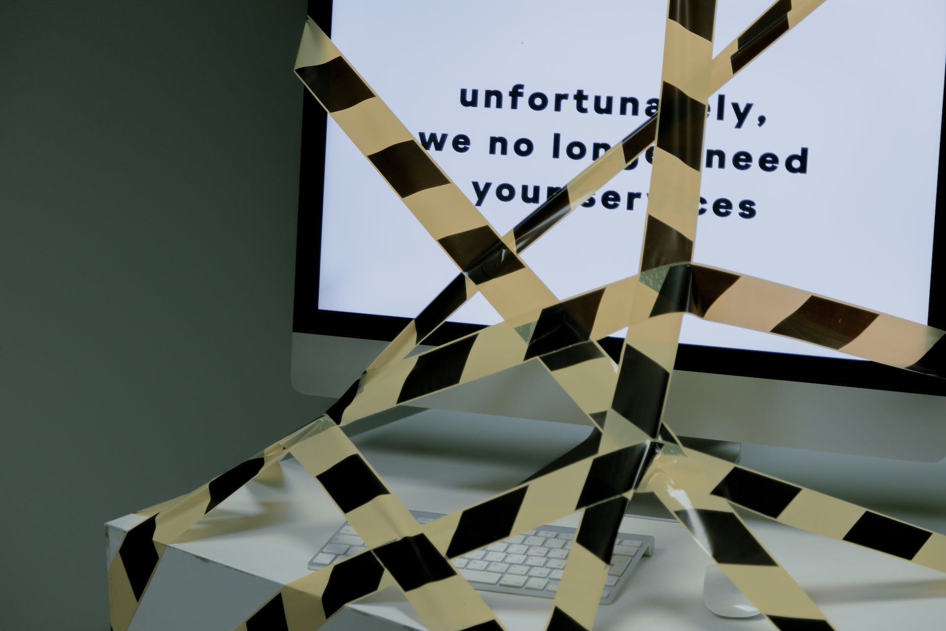 A computer screen displays the text' unfortunately, we no longer need your services'. The screen is on a white desk and the desk has been masked off with black and hells striped hazard tape to stop people using the keyboard on the desk
